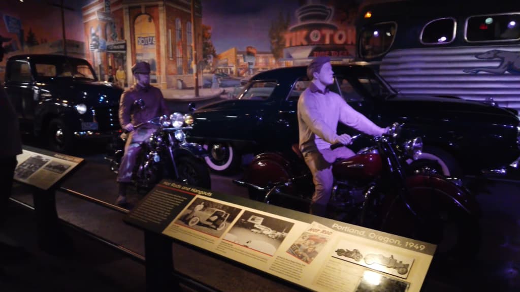 Lifelike statues of 1940s bikers with motorcycles next to vintage cars